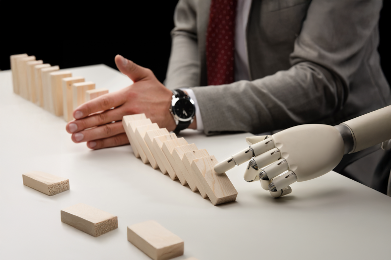 Emberi és robot kéz közösen dominósort indít el, amelyet az ember egy ponton megállít – a stratégiai kontroll és a vezetői vakság szimbolikus ábrázolása az AI korszakában.